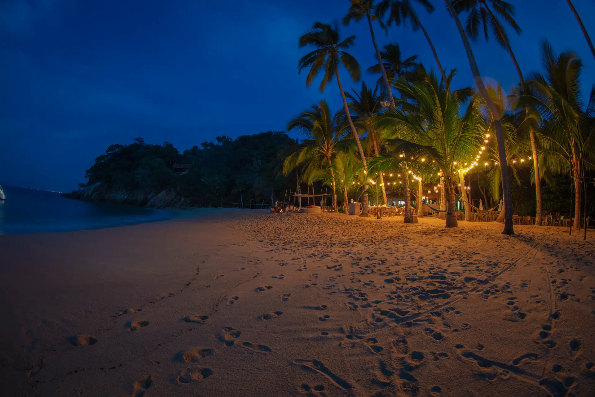 Majahuitas beach at dusk, string lights strung between the palms above the sand, with the cove and the Sierra Madre beyond